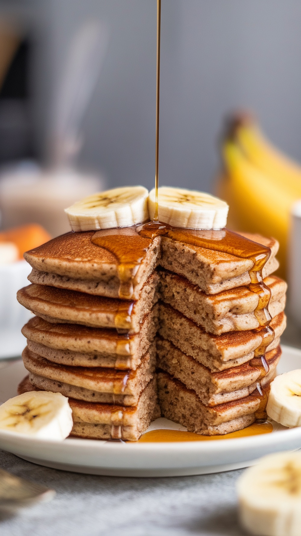 A stack of whole grain banana pancakes topped with banana slices and drizzled with syrup.